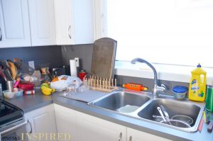 White kitchen cabinets, gray counter top, dark gray tile backsplash, metal sink filled with dishes. A sunny window is over the sink. The counters are covered with clutter - dish rack with a cookie sheet, bananas, grocery bag, coffee supplies, paper towel, a glass tiered desert dish filled with random items.