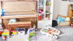 A chaotic disaster of a living room. Toys for young children cover the gray carpet on the bottom third of the photo. A large mid-tone wood piano and white cube bookcase fill the left side of the wall. A black blocks bin and dress up box are low to the ground on the right with a gray flower area rug under them. 